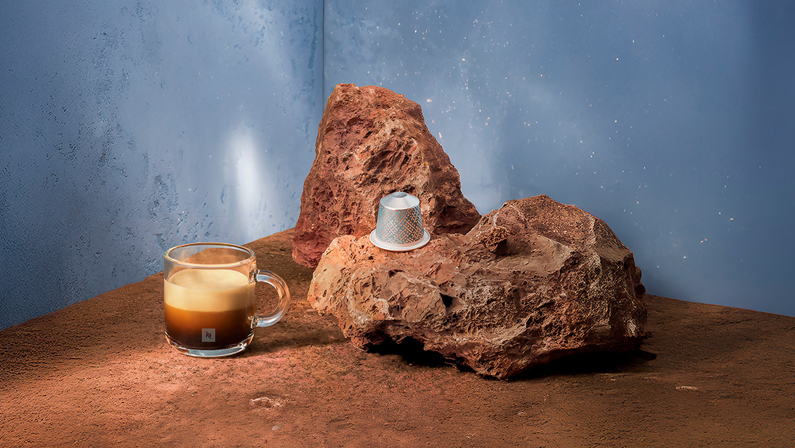 Bodegón encima piedras y arena rojizas con cápsula y taza con café de Nespresso