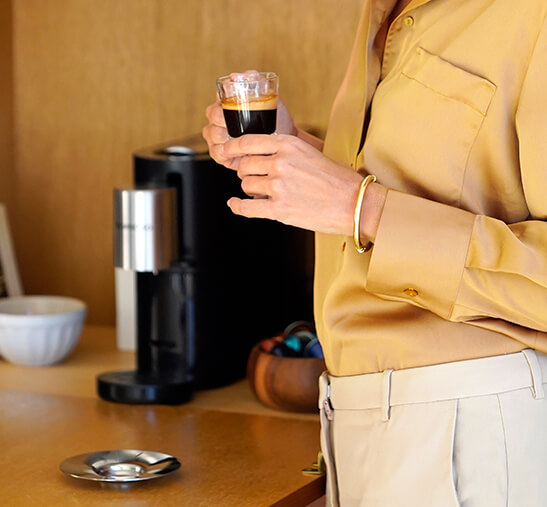 Mujer tomando café Nespresso y máquina Nespresso Vertuo de fondo