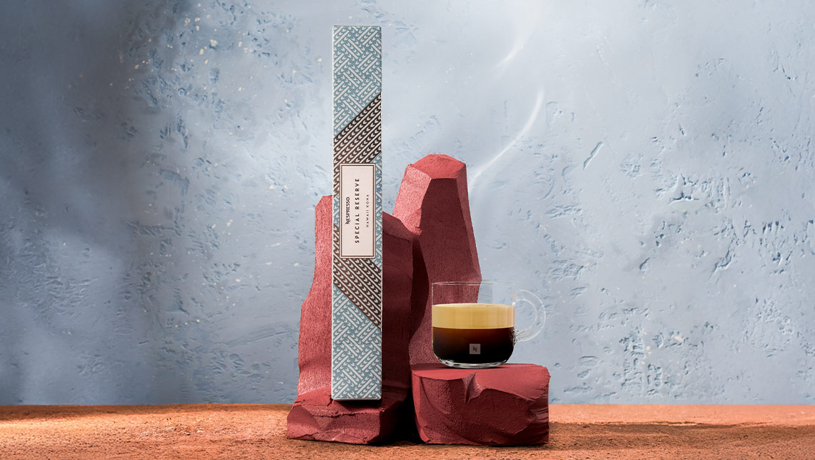 Caja y taza de café Hawaii Kona en un bodegón de piedra y arena rojiza con fondo de pared de piedra azul