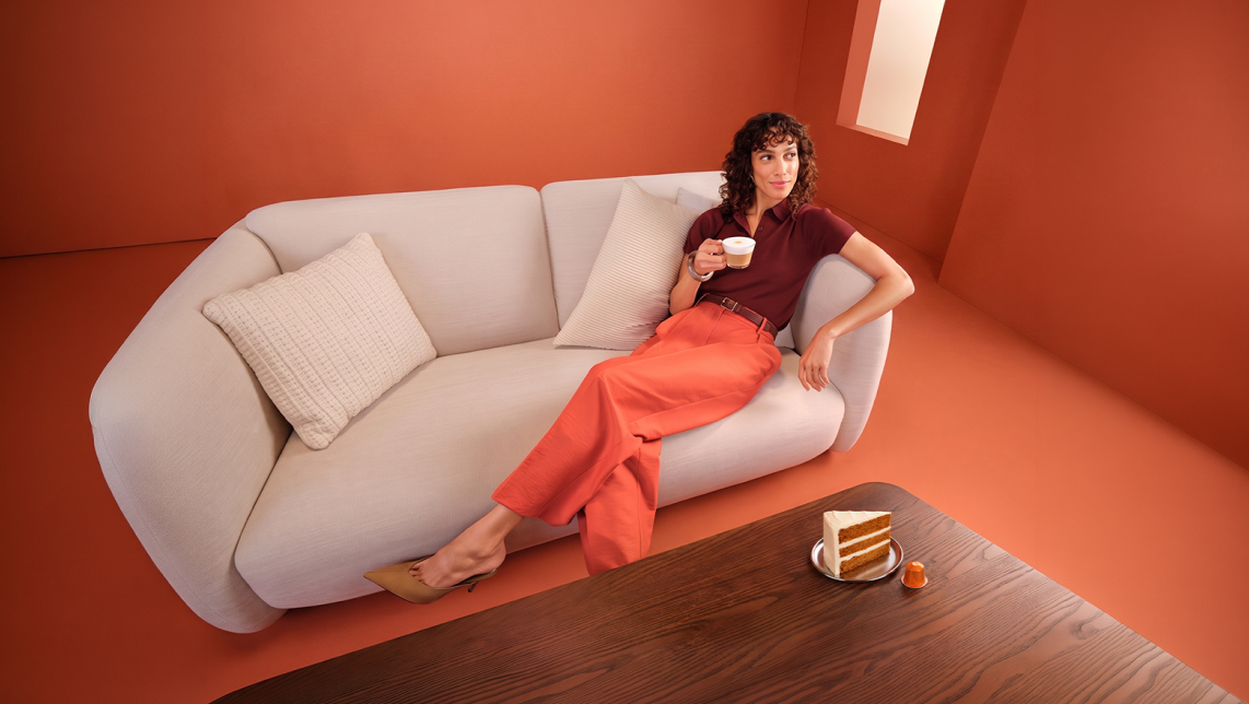 Mujer sentada en un sofá gris ubicado en una habitación naranja junto a su café y tarta pumpkin