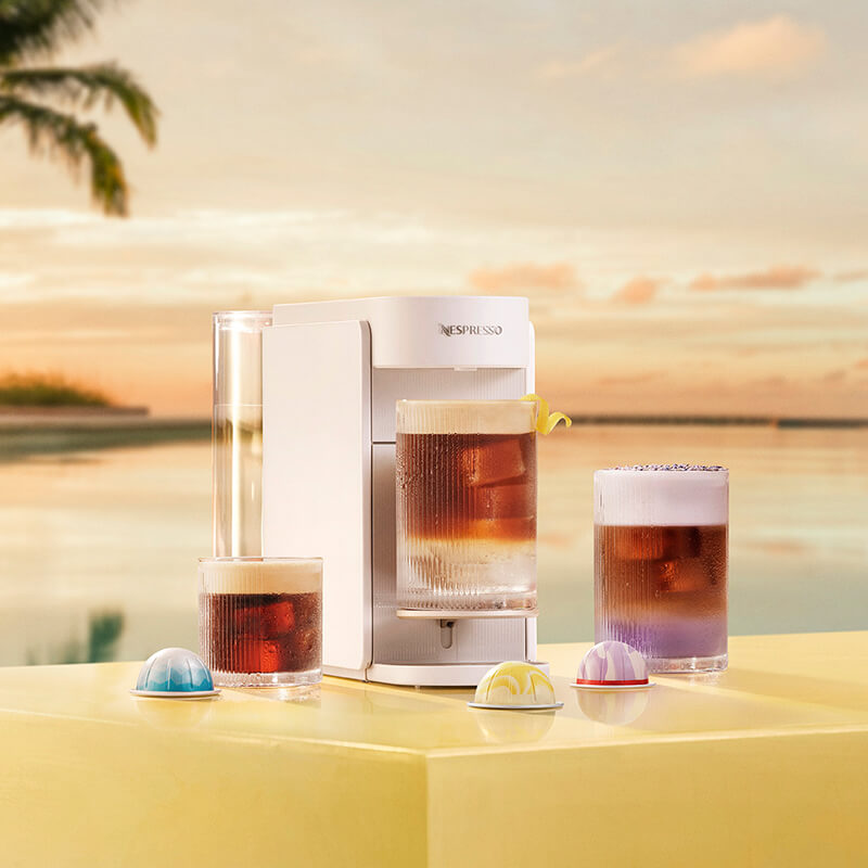 Trois cafés glacés à côté et sous une machine Nespresso Vertuo blanche, avec des capsules posées sur un comptoir jaune, sur fond de plage ensoleillée.