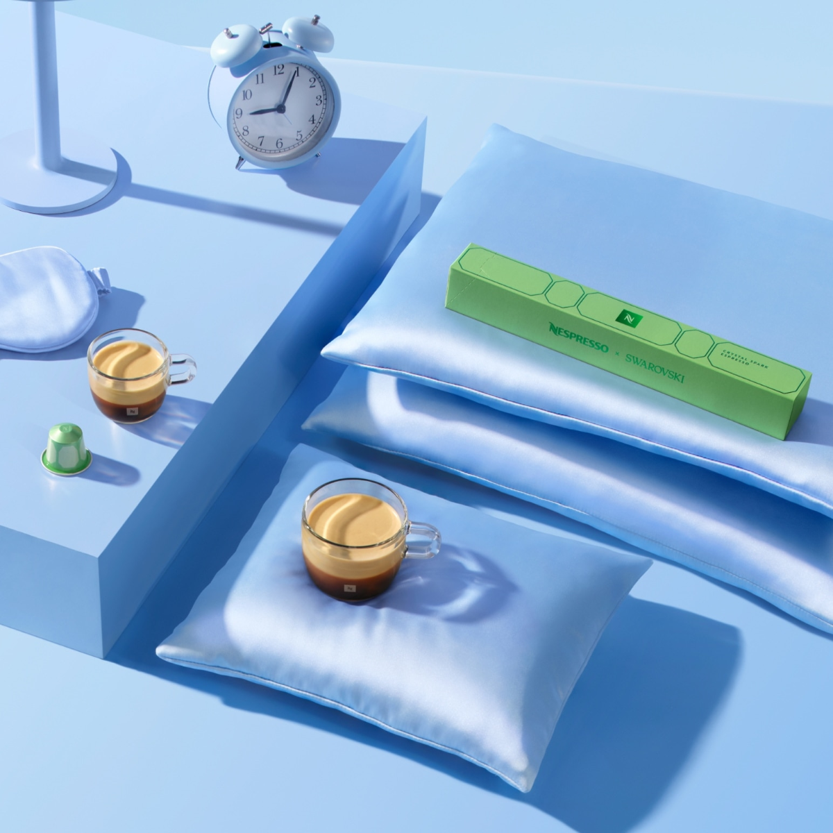 A green Crystal Spark sleeve on blue silk pillows, with two glass cups of espresso, a green Original capsule, an alarm clock and a sleep mask