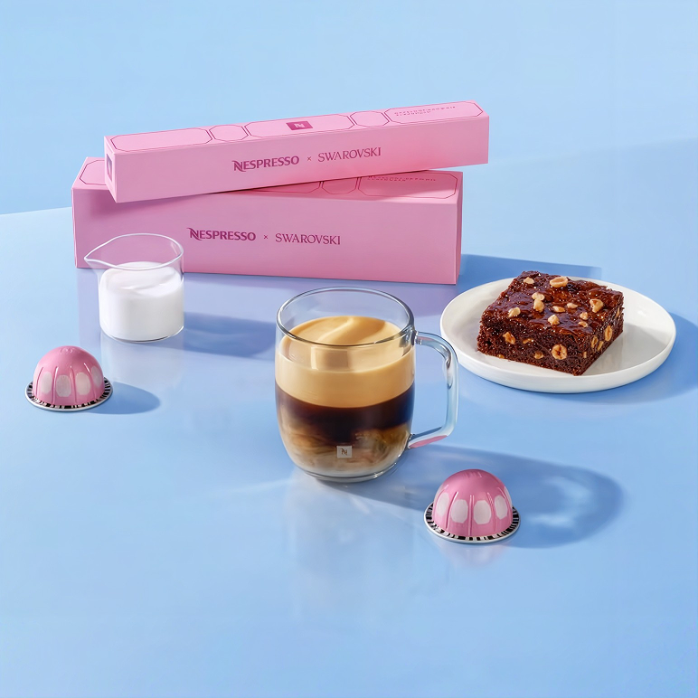 Two Hazelnut Brownie sleeves beside a glass mug of coffee, a jug of milk, a brownie and two pink Vertuo capsules