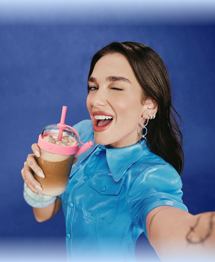 Dua Lipa holding a pink iced coffee drink against a bright blue background