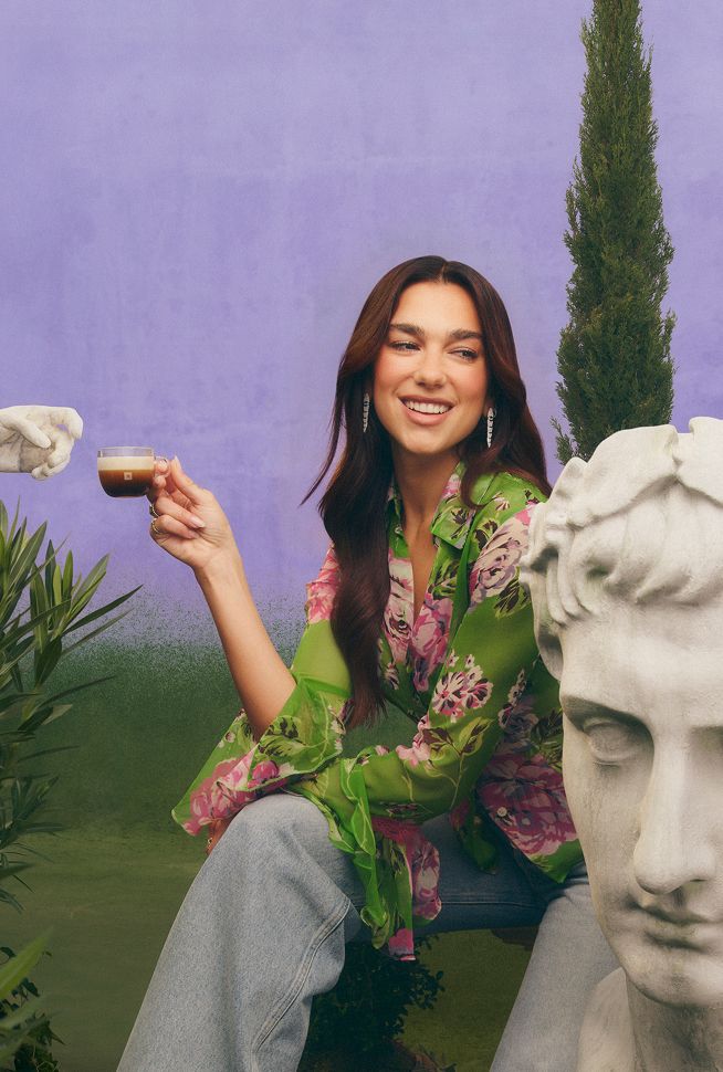 Dua Lipa holding a coffee cup outdoors beside a large classical stone sculpture and greenery.