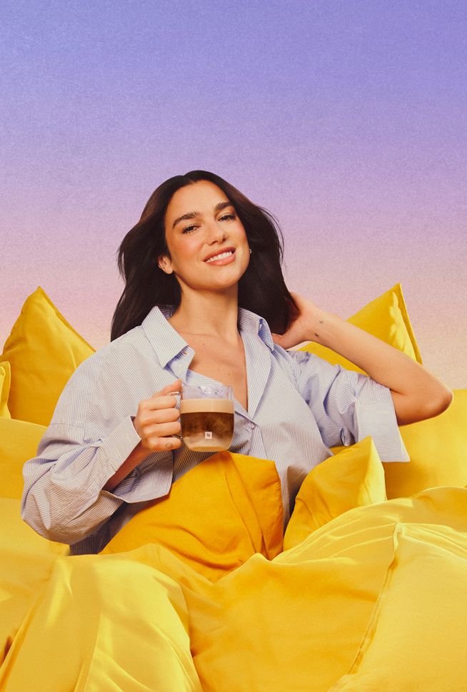 Dua Lipa sitting on vibrant yellow fabric while holding a glass coffee cup