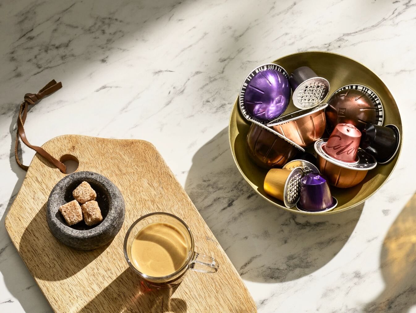 Decorative image of Nespresso pods in a gold bowl next to a black Nespresso recycling bag on a white marble counter.