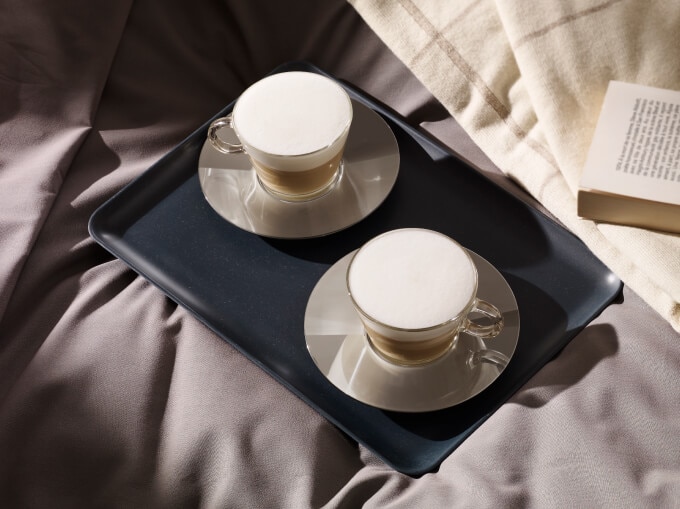 Deux cappuccinos sur plateau sur un lit près d’un livre, parfait pour une matinée confortable.