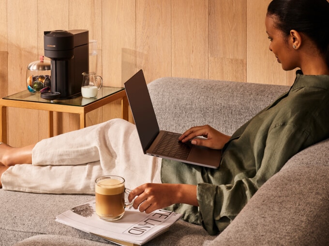 Femme sur canapé avec ordi et café sur journal pour éviter taches, machine Nespresso en arrière-plan.