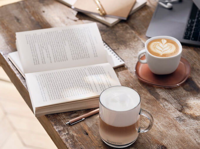 Two lattes, one with beautiful coffee art, sit on a wooden table next to an open book, laptop, and notepad. 