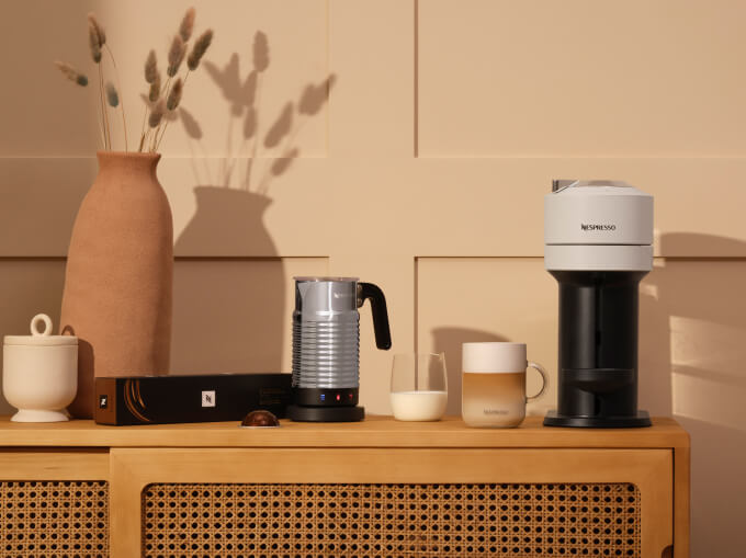 Machine Vertuo Pop blanche et Aerocino sur une console en rotin avec capsule café, lait, latte délicieux et deux poteries en argile.